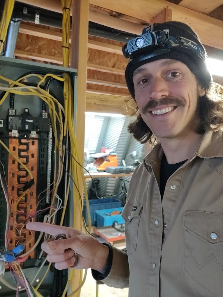 Aaron Clark, owner of Light The Way Electrical, smiling and pointing to wiring inside an open electrical panel